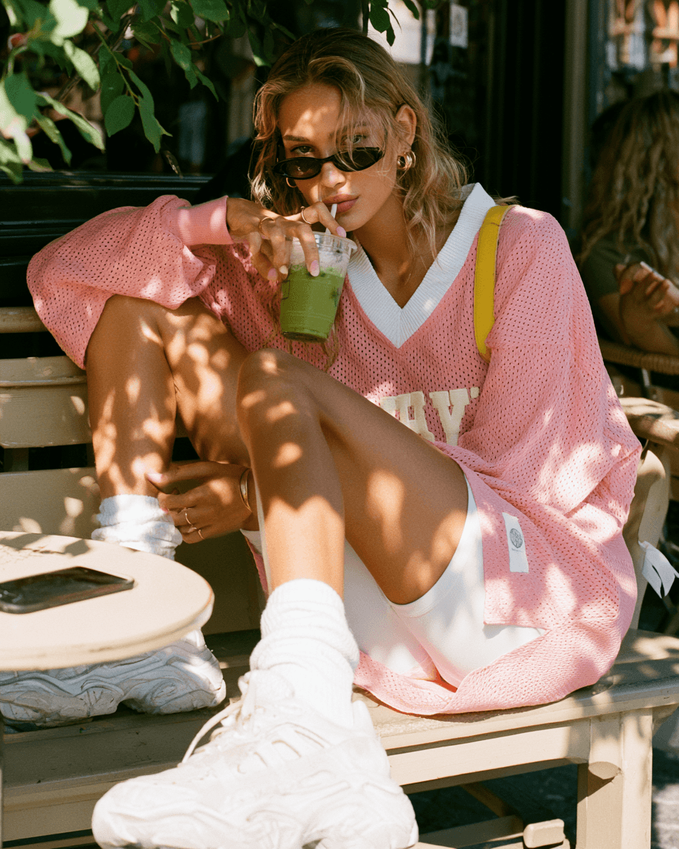One woman sitting sideways on a café terrace bench with one leg up, holding an iced matcha, phone face down on the table. Blush oversized jersey worn as a dress, chunky white trainers, sunglasses. Dappled midday sun through leaves.